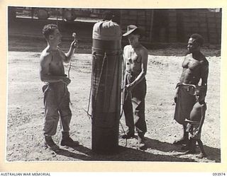 PALMALMAL PLANTATION, JACQUINOT BAY, NEW BRITAIN, 1945-07-13. A NATIVE AND HIS PICCANINNY WATCHING PRIVATE G. WHEELER (1) AND PRIVATE K. WILSON (2), MEMBERS OF HEADQUARTERS 31 SUPPLY DEPOT COMPANY ..