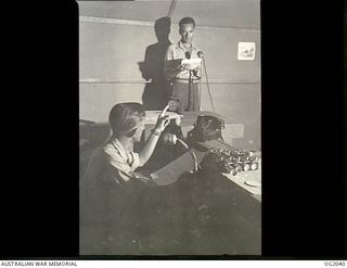 MADANG, NEW GUINEA. C. 1944. LEADING AIRCRAFTMAN K. D. ROBINSON, WEST MAITLAND, NSW, DUTY ANNOUNCER AT RAAF RADIO MADANG, GIVES THE "TAKE IT AWAY" SIGN TO CORPORAL R. J. POYNTON, GEELONG, VIC, AT ..