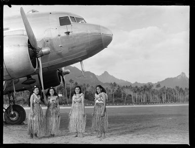 Four unidentified local girls wearing hula skirts in front of a C47 transport aircraft, Rarotonga airfield, Cook Islands