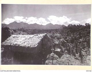 MOROKAIMORO, BOUGAINVILLE, 1945-06-12. A NATIVE HUT AT THE ANGAU CAMP. THE SOUTHERN END OF CROWN PRINCE RANGE IS IN THE BACKGROUND. A COMPANY OF 1 PAPUAN INFANTRY BATTALION TROOPS OPERATES WITH 2/8 ..