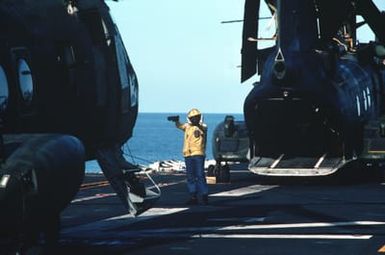 A helicopter launch petty officer directs flight deck operations aboard the amphibious assault ship USS GUAM (LPH 9) during operations off the coast of Lebanon. Two CH-46 Sea Knight helicopters are parked on the flight deck