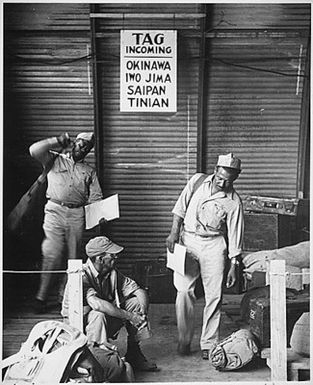 "Two soldiers gather up their baggage as transportation arrives to take them to their outfit on Guam. Another soldier sits disconsolately awaiting further orders of transportation."