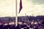 Newly adopted Papua New Guinea flag raised at Erave patrol post, watched by crowd including decorated men and saluting patrol officer
