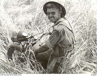 PORT MORESBY, PAPUA. 1942-07-11. A HOT AND DUSTY AUSTRALIAN DESPATCH-RIDER STOPS TO SMILE AT THE CAMERA WHILE TRYING TO FORCE HIS MOTOR CYCLE THROUGH THE LONG GRASS WHILE ENGAGED ON MANOEUVRES IN ..