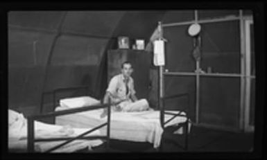 [Serviceman sitting on bed in barracks building]