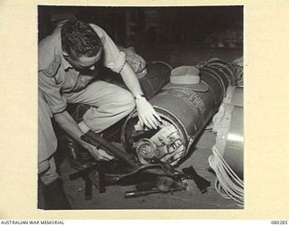 NADZAB, NEW GUINEA. 1944-10-21. VX108086 CAPTAIN W.B. BALLARD, AT HEADQUARTERS NEW GUINEA FORCE EXAMINING OWEN GUNS AND OTHER STORES DROPPED BY PARACHUTE IN A STORPEDO CONTAINER