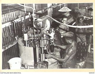 LAE, NEW GUINEA. 1944-12-12. CORPORAL A.P. RYAN (1), SUPERVISES NATIVES SUPPLIED BY ANGAU AS THEY OPERATE A FILLING AND CAPPING MACHINE FOR ARMY CANTEENS SERVICE AT A SOFT DRINK FACTORY WITHIN LAE ..