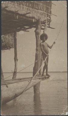 Man standing at the prow of a canoe with a house on stilts behind, Hanuabada village, Papua, 1921 / Sarah Chinnery