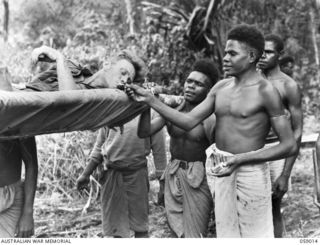 FARIA VALLEY, NEW GUINEA. 1943-10-16. SX17161 PRIVATE L. G. AGARS IS GIVEN A LIGHT FOR HIS CIGARETTE BY A NATIVE WHILE BEING EVACUATED FROM GUY'S POST TO THE MAIN DRESSING STATION