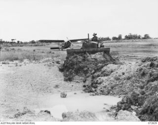 MADANG, NEW GUINEA, 1944-04-30. NX99280 CORPORAL FRANKHEISS, ROYAL AUSTRALIAN ENGINEERS USES A BULLDOZER TO CLEAR THE MADANG AIRFIELD. A STINSON AIRCRAFT IS SEEN AT THE BACKGROUND