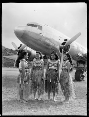 Four unidentified local girls wearing hula skirts in front of a C47 transport aircraft, Rarotonga airfield, Cook Islands