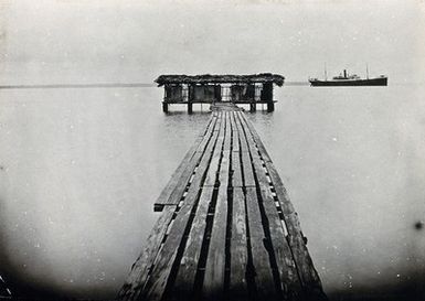 ["Buratu, Fiji (?): latrine buildings on a river, at the end of a long wooden jetty, with a steamer boat seen in the background. Photograph, 1910/1920."]