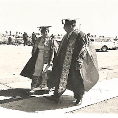 Photo of male and female dressed in graduation gowns; male walking over woven pandanus mat (67534)