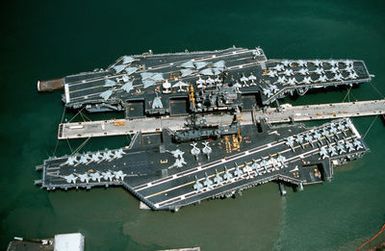 An aerial view of various aircraft lining the flight decks of the aircraft carrier USS INDEPENDENCE (CV-62), right, and USS MIDWAY (CV-41) moored beside each other in the background. The MIDWAY is en route from Naval Station, Yokosuka, Japan, to Naval Air Station, North Island, California, where it will be decommissioned in the spring of 1992. The INDEPENDENCE will travel to Yokosuka to take over as the Navy's forward-based aircraft carrier