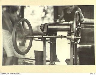 KOKODA, NEW GUINEA. 1944-04-11. NATIVES AT A RUBBER PLANTATION FEEDING RUBBER THROUGH A PATTERN ROLLER WHICH IMPRESSES THE TRADE MARK. THIS PROCESS COMPLETES THE FINAL REMOVAL OF EXCESS WATER FROM ..