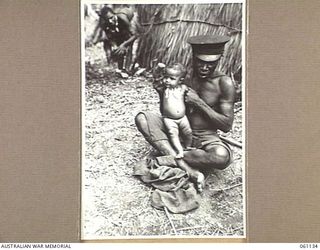 DUMPU AIRSTRIP, NEW GUINEA. 1943-11-14. A POLICE BOY NURSING A NATIVE BABY WHILE THE MOTHER WORKS IN THE 7TH AUSTRALIAN DIVISION HEADQUARTERS AREA