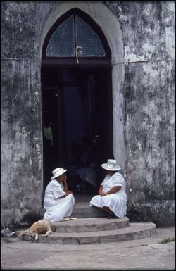 Women and a dog resting on steps outside church, Rarotonga