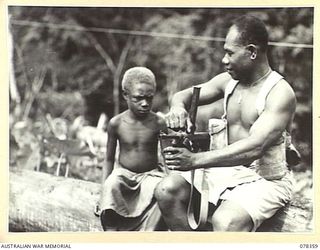 TAMKAIDAN, NEW BRITAIN. 1945-01-14. SERGEANT MARTIN POIRIRI, VETERAN ALLIED INTELLIGENCE BUREAU SOLDIER SHOWING A YOUNG NATIVE LAD FROM A NEARBY VILLAGE HIS OWEN GUN