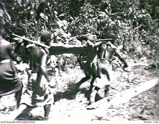 THE SOLOMON ISLANDS, 1945-04-24/27. FOUR NATIVE PORTERS CARRYING A PATIENT ON A BUSH STRETCHER ALONG A TRACK ON BOUGAINVILLE ISLAND. (RNZAF OFFICIAL PHOTOGRAPH.)