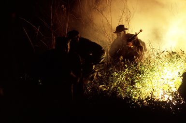 Two Marines move past a burning flare as they advance on a Seabee camp during exercise Kennel Bear '89. Marines from Marine Barracks Guam are acting as aggressors against Seabees from Naval Mobile Construction Battalion 3 (NMCB-3) and Marines from the 3rd Force Service Support Group (3rd FSSG)