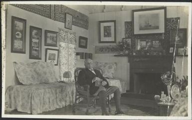 Gentleman wearing riding clothes seated in his wife's sitting room which is decorated in the English style but includes tapa cloth wall hangings, Fiji, 1918