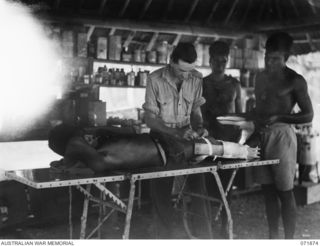 LAE, NEW GUINEA. 1944-03-30. LEFT TO RIGHT, STANDING: QX56754 CORPORAL G. C. HARRIS, ASSISTED BY MEDICAL ORDERLY MUNTAM, AND CORPORAL YASIA, BANDAGES THE BROKEN LEG OF PIRUNGA (PATIENT), AT THE ..