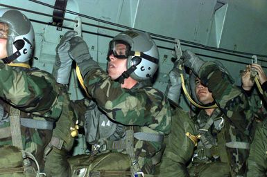 Left side front view medium close-up of US Marines from 2nd Platoon, Company A, 5th Reconnaissance Battalion, 3rd Marine Division, as they attach the static lines to the cable from their MC1-1 parachutes before jumping over the drop zone from a USAF C-141 Starlifter during Force Reconnaissance Exercises