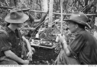 1942-12-04. NEW GUINEA. GONA. TROOPS SNATCH A HASTY MEAL DURING A LULL IN THE GONA FIGHTING. IN BACKGROUND IS A JAPANESE GRAVE. THIS PHOTO WAS TAKEN LESS THAN 100 YARDS FROM THE JAPANESE POSITIONS. ..