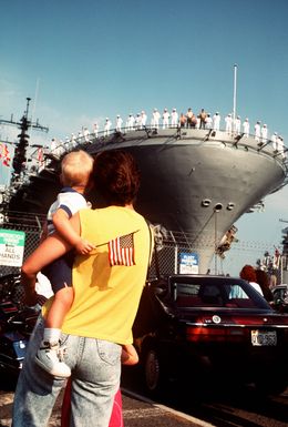 A little boy watches from his mother's arms as his father departs for the Persian Gulf aboard the amphibious assault sh USS GUAM (LPH-9), one of several ships deploying from Norfolk in response to Iraq's invasion of Kuwait