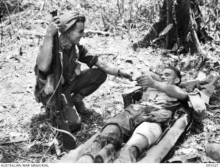 BOUGAINVILLE. 1945-04-17. PRIVATE R.R. COOK, 24 INFANTRY BATTALION (2), SHOWING A TOBACCO TIN THROUGH WHICH SHRAPNEL FROM A GRENADE ENTERED HIS LEG, TO HIS FRIEND PRIVATE H. WASHUSEN (1). HE ..