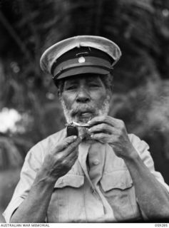 HOPOI, NEW GUINEA, 1943-10-30. PORTRAIT OF DAHU, PARAMOUNT CHIEF OF THE HOPOI DISTRICT, LIGHTING HIS PIPE WITH JAPANESE INVASION MONEY TO DEMONSTRATE TO HIS FELLOW TRIBESMEN ITS LACK OF VALUE