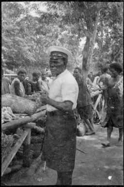 Man wearing an officer style cap standing beside a trestle table of produce at the  Boong, the native market, Rabaul, New Guinea, ca. 1929 / Sarah Chinnery