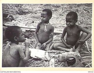 KIARIVU, NEW GUINEA, 1945-08-11. THREE NATIVE BOYS AGED THREE YEARS GROUPED ROUND THEIR SMALL FIRE SMOKING. TROOPS OF 2/7 INFANTRY BATTALION ARE NOW IN THE AREA IN CONTROL OF THE EMERGENCY LANDING ..