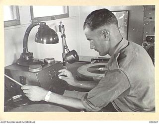 WEWAK, NEW GUINEA. 1945-10-29. SERGEANT N.S. BRAY, 1 BROADCASTING STATION, AUSTRALIAN ARMY AMENITIES SERVICE, AT THE MICROPHONE OF RADIO STATION 9AJ SOUNDING THE BROADCASTING GONG