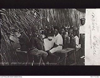 NEW BRITAIN. C. 1914. NATIVE POLICE AT THEIR MEAL TABLE AT MATUPI, NEAR RABAUL. THESE MEN WERE EMPLOYED BY THE GERMAN AUTHORITIES IN NEW GUINEA. NOTE GERMAN HATS. (DONOR: J. TONGS)