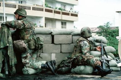 Two Marines man a security post on the roof of the US Embassy. The Marine at right is armed with an M16A2 rifle; the Marine at left is leaning on an AT4 light anti-armor weapon covered with a poncho. Marines of the 22nd Marine Expeditionary Unit (22nd MEU), deployed aboard the amphibious assault ship USS SAIPAN (LHA 2), were sent to augment security at the embassy as part of Operation SHARP EDGE