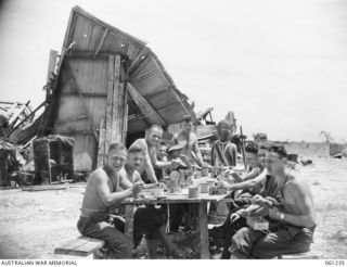 SATTELBERG, NEW GUINEA. 1943-11-29. TROOPS OF A COMPANY, 2/48TH AUSTRALIAN INFANTRY BATTALION ENJOYING THEIR LUNCH. THE BOMBED BUILDING IN THE BACKGROUND IS THE LUTHERAN CHURCH, AND THE FIGURE AT ..