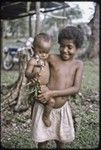 Girl holds infant wearing short red shell necklace, infant has stick with a butterfly on it