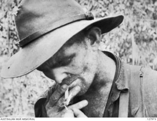 NAMLING, NEW GUINEA. 1943-07-19. PRIVATE TOM KIDD, A MEMBER OF 2/3RD AUSTRALIAN INDEPENDENT COMPANY, RECEIVING A LIGHT FOR HIS CIGARETTE AFTER HIS UNIT'S RETURN FROM ACTION IN THE "GRAVEYARD" AREA, ..