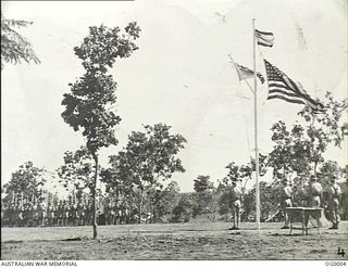 BUNA, NEW GUINEA. C. 1943. TROPICAL PARADE GROUND OVER WHICH FLY THE STAR SPANGLED BANNER, THE FLAG OF THE RAAF AND THE SQUADRON LEADER'S FLAG WITH RAAF TROOPS FORMED UP AWAITING THE DECORATION OF ..