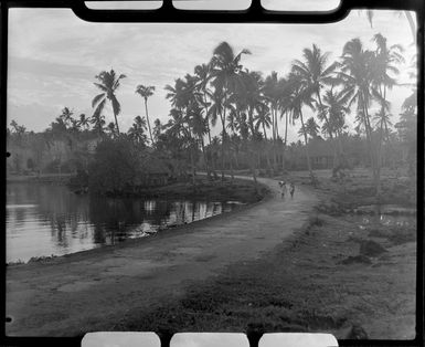 Two unidentified girls walking down road at Faleolo, Apia, Upolu, Samoa, palm trees and fales in background