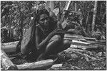 Woman with netbag holds infant, firewood stacked nearby