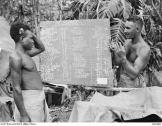DUMPU, NEW GUINEA. 1943-11-13. WX6512 DRIVER B. O'DONOHUE OF HEADQUARTERS, 7TH AUSTRALIAN DIVISION ACTING AS A BOOKMAKER FOR THE MELBOURNE CUP, TRYING TO EXPLAIN THE CUP TO A NATIVE BOY