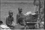 Mortuary ceremony: mourning women, faces and bodies painted black, beside a basket filed with banana leaf bundles