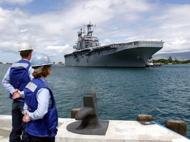 Two US Navy (USN) Sailors standby to tie up a ship at a mooring bitt on a pier at Naval Station Pearl Harbor. In the background the USN Tarawa Class Amphibious Assault Ship USS TARAWA (LHA 1) Sailors "man the rails" as the ship pulls into the harbor. The TARAWA is in Pearl Harbor to take part in Rim of the Pacific (RIMPAC) 2004. RIMPAC is the largest international maritime exercise in the waters around the Hawaiian Islands. This years exercise includes seven participating nations; Australia, Canada, Chile, Japan, South Korea, the United Kingdom and the United States. RIMPAC enhances the tactical proficiency of participating units in a wide array of combined operations at sea, while...