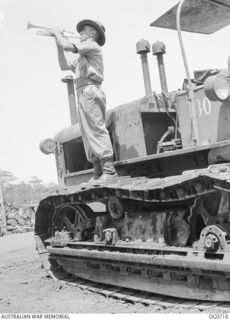 NADZAB, NEW GUINEA. C. 1944-02. 76295 LEADING AIRCRAFTMAN (LAC) CLIFFORD DAVID SEARLE OF EAST BRISBANE, QLD, BLOWING REVEILLE AT NO. 62 MOBILE WORKS SQUADRON RAAF STANDING ON THE TRACKS OF A GIANT ..