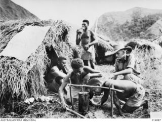 NEW GUINEA. 8 NOVEMBER 1943. MEMBERS OF A PAPUAN INFANTRY UNIT IN CAMP AT A FORWARD POST. LT. A. HALL, OF BARDON, QUEENSLAND, INSPECTS HUTS AND WEAPONS