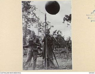 MAMAGOTA AREA, BOUGAINVILLE. 1945-07-22. GUNNER J.R. HAMILTON (2) RELEASING A HYDROGEN FILLED BALLOON TO RECORD WIND SPEED AND DIRECTION FOR ARTILLERY CORRECTIONS. SERGEANT R.M. PARNABY (1) IS ..