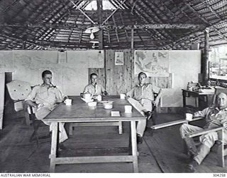 GOODENOUGH ISLAND, NEW GUINEA. 1944-05. OFFICERS TAKE TEA IN THE MAP LINED WARDROOM. LEFT TO RIGHT: SUB LIEUTENANT B. Y. HALL, LIEUTENANT H. HARVIE, LIEUTENANT A BUSH AND LIEUTENANT COMMANDER S. E. ..