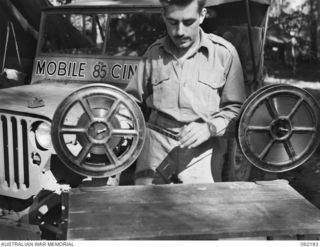 DUMPU, NEW GUINEA. 1943-12-10. VX145795 LANCE SERGEANT E. W. FRANKLIN, ASSISTANT OPERATOR OF THE 85TH AUSTRALIAN MOBILE CINEMA UNIT, AUSTRALIAN ARMY AMENITIES, 7TH AUSTRALIAN DIVISION, WINDING FILM ..